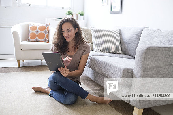 Junge Frau sitzt auf dem Boden im Wohnzimmer und benutzt ein digitales Tablet