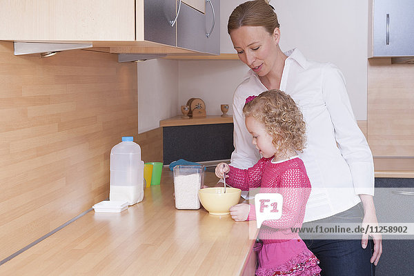 Mutter beobachtet Tochter (4-5) beim Mischen von Zutaten in einer Schüssel
