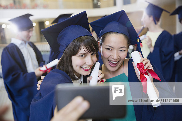 Lächelnde weibliche Hochschulabsolventinnen in Mütze und Kittel  die Selfie nehmen.