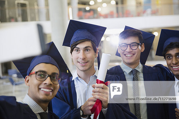 Male college graduates in cap and gown with diploma taking selfie