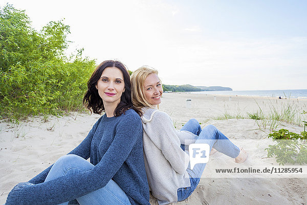Two women on sandy beach sitting back to back