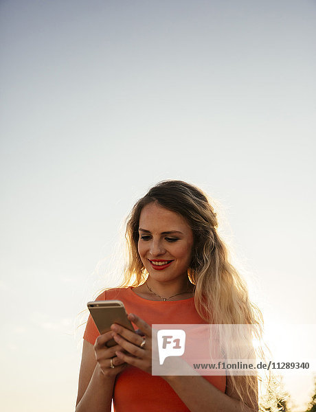 Lächelnde blonde Frau blickt auf das Smartphone vor dem Himmel