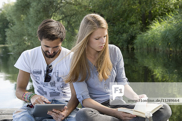 Young couple sitting on jetty at a lake with book and tablet