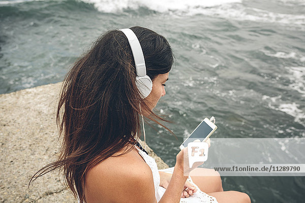 Frau hört Musik mit Kopfhörern und schaut auf das Smartphone vor dem Meer.