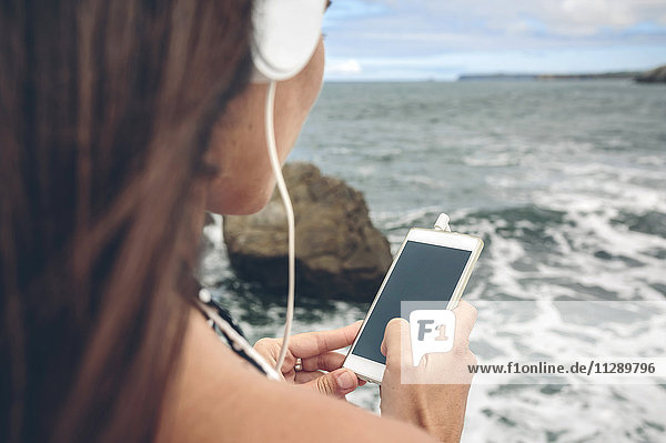 Woman using smartphone while listening with headphones in front of the sea