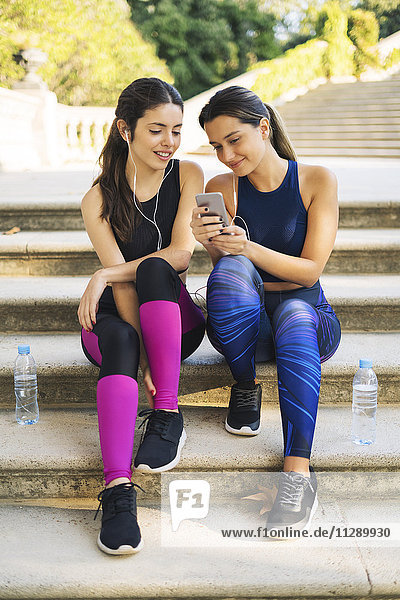 Zwei sportliche junge Frauen sitzen auf der Treppe und schauen aufs Handy.
