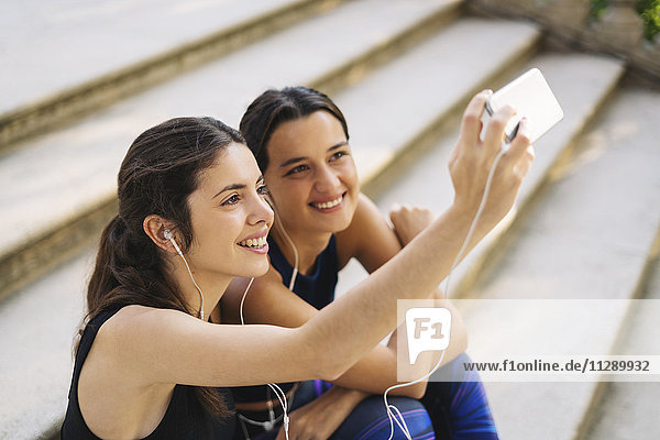 Zwei sportliche junge Frauen sitzen auf einer Treppe und nehmen einen Selfie.