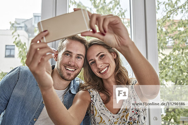 Porträt eines lächelnden Paares vor dem Fenster  das Selfie mit Smartphone nimmt