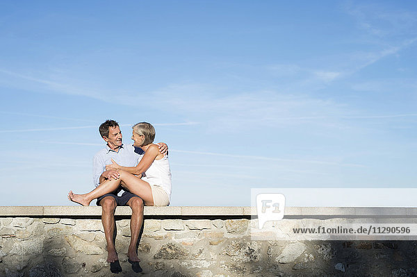 Senior couple sitting barefoot on a wall in front of sky