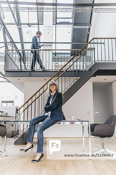 Businesswoman sitting on desk in office with businessman walking on upper floor