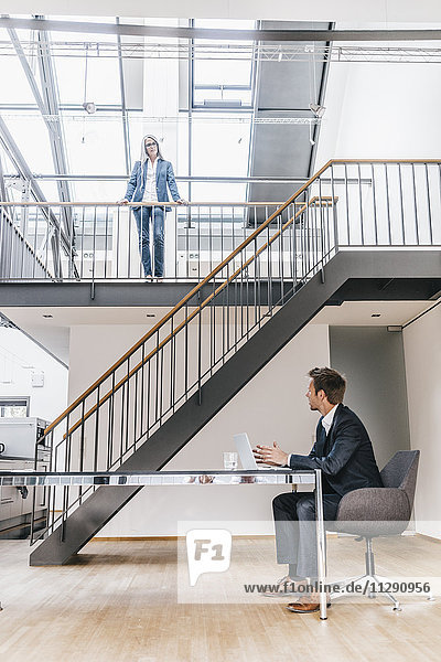 Businessman sitting at desk in office with businesswoman on upper floor