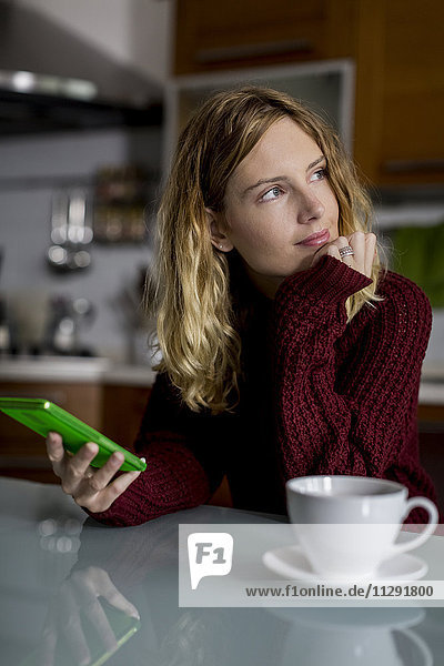 Nachdenkliche Frau sitzend in der Küche mit Smartphone und Tasse Tee