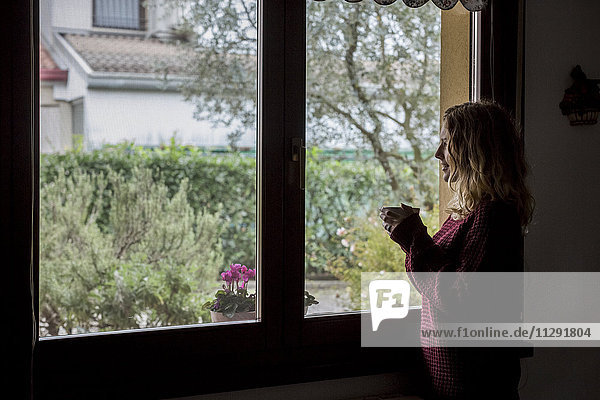 Frau mit Tasse Tee durchs Fenster schauend