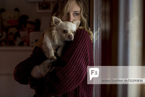 Frau schaut durchs Fenster mit Hund auf den Armen