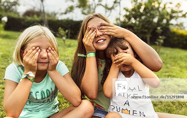 Drei Mädchen  die die Augen mit den Händen auf einer Wiese bedecken.