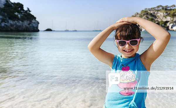 Porträt eines lächelnden kleinen Mädchens mit Sonnenbrille vor dem Meer stehend