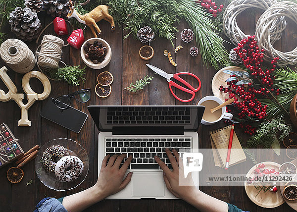 Frau arbeitet mit Laptop an ihrem Schreibtisch  bedeckt mit Utensilien zum Erstellen von Adventskränzen  Teilansicht