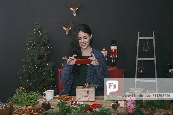 Lächelnde junge Frau fotografiert geschmücktes Weihnachtsgeschenk mit Smartphone