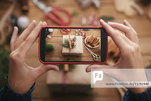 Frau fotografiert dekoriertes Weihnachtsgeschenk mit Smartphone  Nahaufnahme