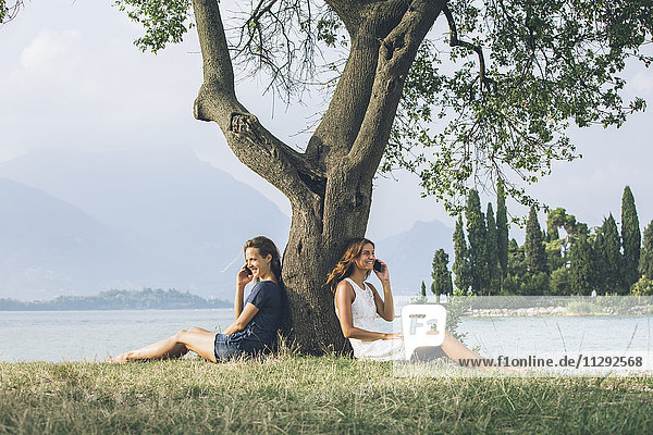Italy  Lake Garda  two young women leaning against a tree talking on cell phones