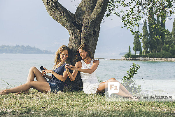 Italien  Gardasee  zwei junge Frauen  die sich mit dem Handy an einen Baum lehnen.