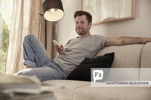 Entspannter Mann zu Hause sitzend auf der Couch mit Handy