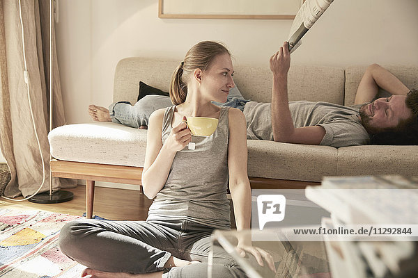 Frau trinkt Tee im Wohnzimmer mit Mann auf der Couch liegend mit Zeitung