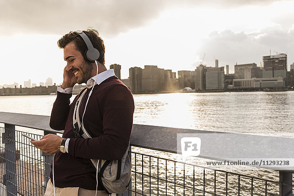 USA  New York City  lächelnder junger Mann mit Kopfhörer und Handy am East River