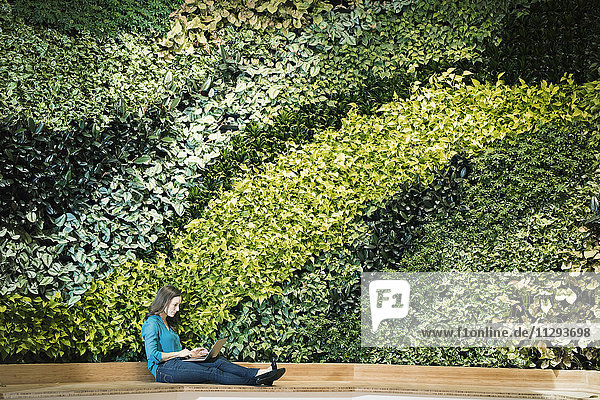 Junge Frau mit Laptop vor grüner Pflanzenwand