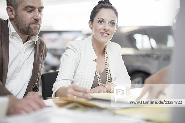 Car dealer and couple sitting at desk with agreement forms in showroom