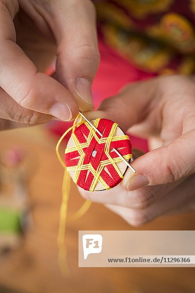 Knopfmacherin  Posamenten Knopf-Rohling aus Holz mit Haltewerkzeug  rotes Sternmuster wird zusätzlich mit gelbem Garn dreifach von Händen umwickelt und genäht  fünfter Schritt