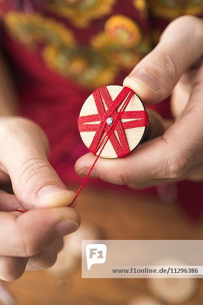 Knopfmacherin  Posamenten Knopf-Rohling aus Holz mit Haltewerkzeug wird mit rotem Garn von Händen umwickelt in Sternmuster  dritter Schritt