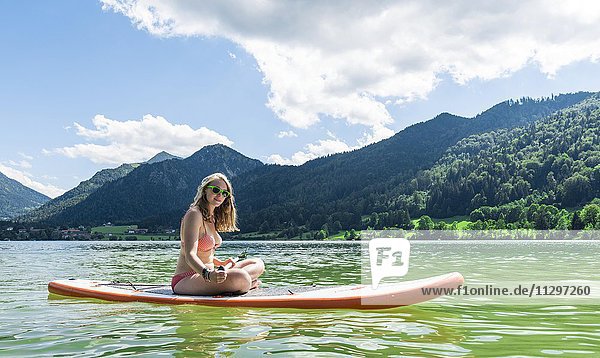 Junge Frau sitzt auf einem Stehpaddel  Stand-Up-Paddel oder SUP  hinten Berge  Schliersee  Oberbayern  Bayern  Deutschland  Europa