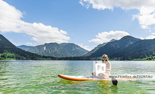 Junge Frau sitzt auf einem Stehpaddel  Stand-Up-Paddel oder SUP  hinten Berge  Schliersee  Oberbayern  Bayern  Deutschland  Europa