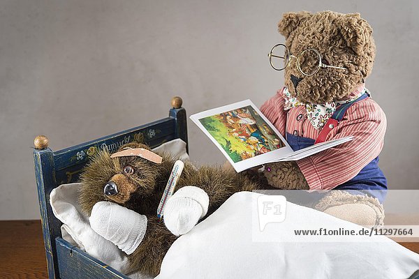Kranker Teddybär  Pfoten verbunden  Pflaster auf Kopf  Fieberthermometer  liegt in Original Pumuckl Bettchen  grosser Bär mit Brille liest aus Buch vor