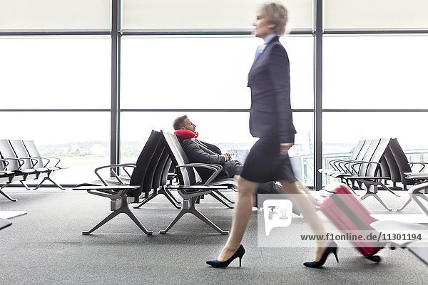 Businesswoman pulling suitcase past resting businessman with neck pillow in airport departure area