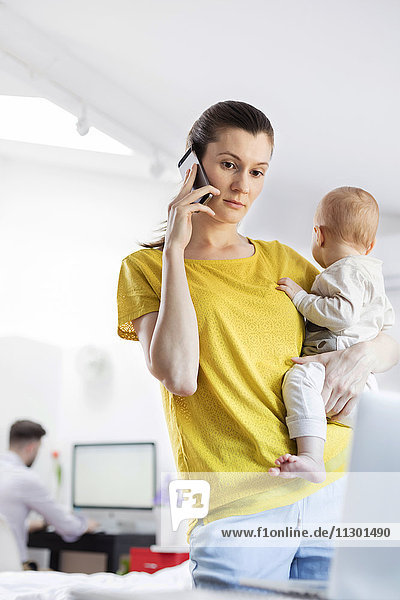 Mutter hält Baby-Tochter und spricht auf dem Handy am Laptop