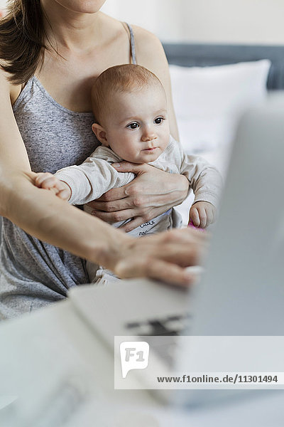 Mutter hält Baby-Tochter und arbeitet am Laptop
