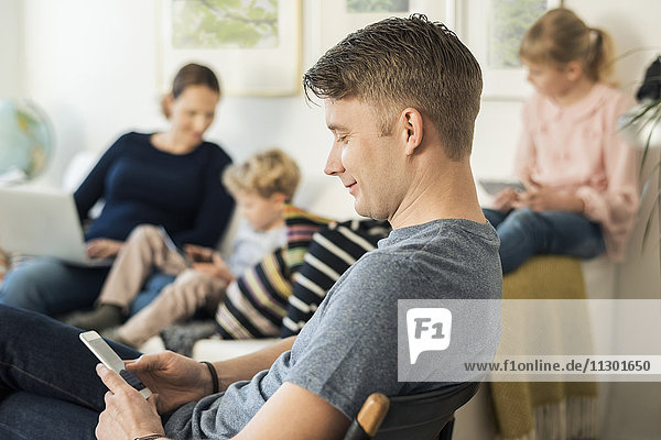 Side view of man using phone with family sitting on sofa at home