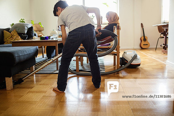 Boy cleaning floor with vacuum cleaner while mother sitting on armchair at home