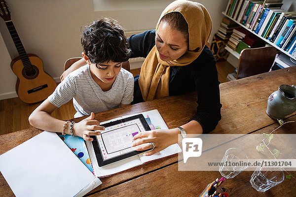 Hochwinkelansicht der Mutter  die den Sohn bei der Verwendung des digitalen Tabletts während des Studiums zu Hause unterstützt.