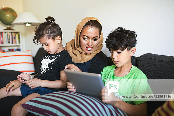 Mutter assistiert dem Sohn bei der Benutzung des digitalen Tabletts  während er zu Hause auf dem Sofa sitzt.