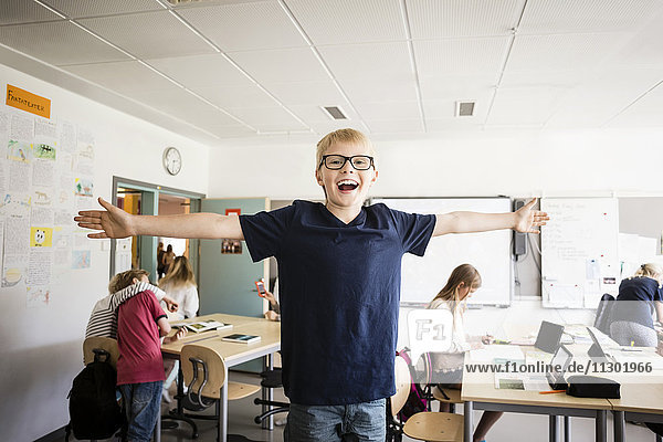 Happy boy standing with arms outstretched against friends in classroom