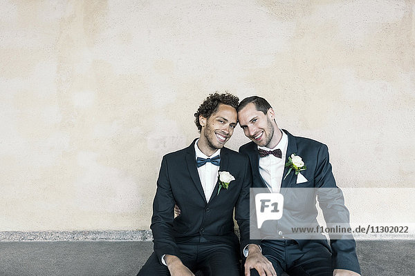 Portrait of newlywed gay couple sitting on bench against wall