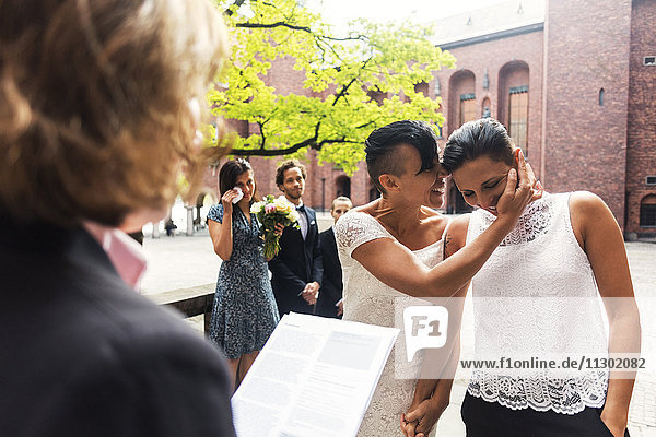 Gäste und Priester suchen fröhliche lesbische Paar flüstern während der Hochzeitszeremonie