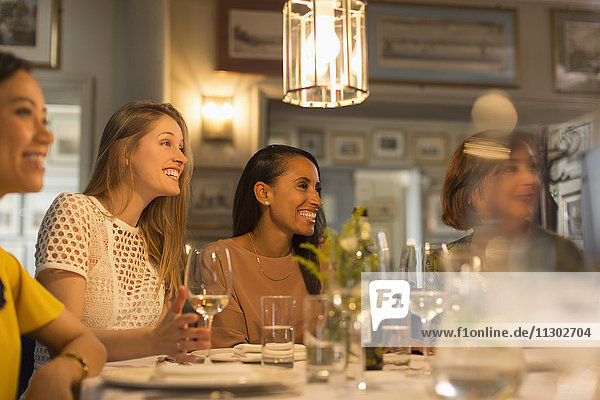Smiling women friends looking away dining and drinking white wine at restaurant table