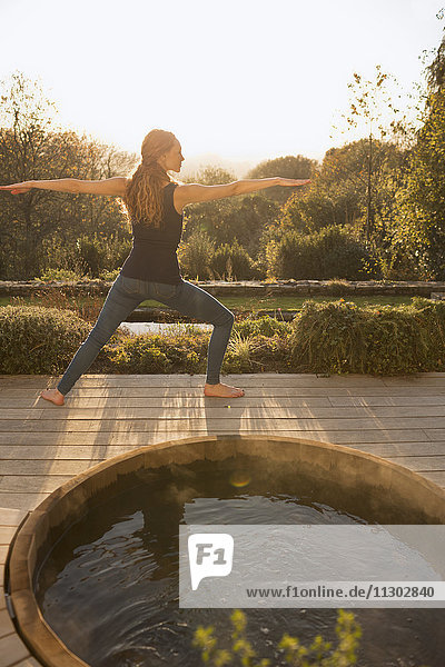 Frau übt Yoga-Krieger 2-Pose auf Herbst-Terrasse mit Whirlpool