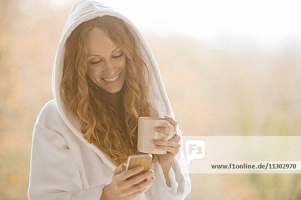 Lächelnde Frau im Kapuzenbademantel  die mit ihrem Handy SMS schreibt und Kaffee trinkt