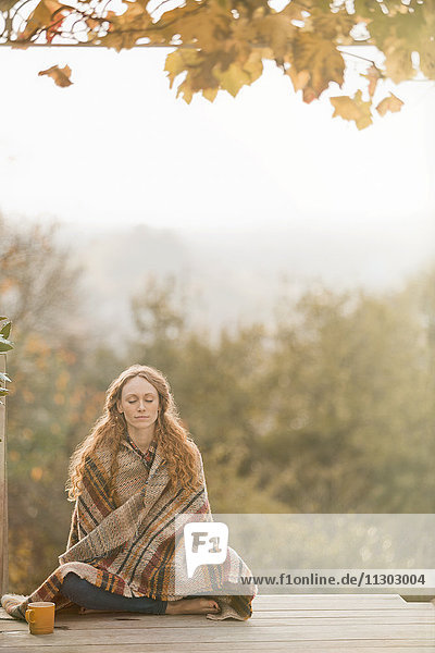 Gelassene Frau  eingewickelt in eine Decke  meditiert auf einer Herbstterrasse