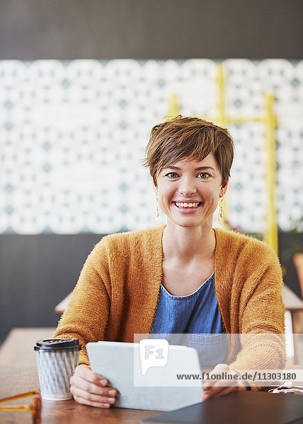 Porträt einer lächelnden Geschäftsfrau  die ein digitales Tablet benutzt und am Kaffeetisch Kaffee trinkt